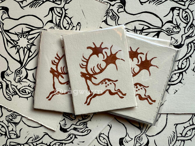 3 small lino prints of a megaloceros (Irish Elk) in a cave-painting-inspired style. Brown ink. The prints are laying on top of multiple other prints